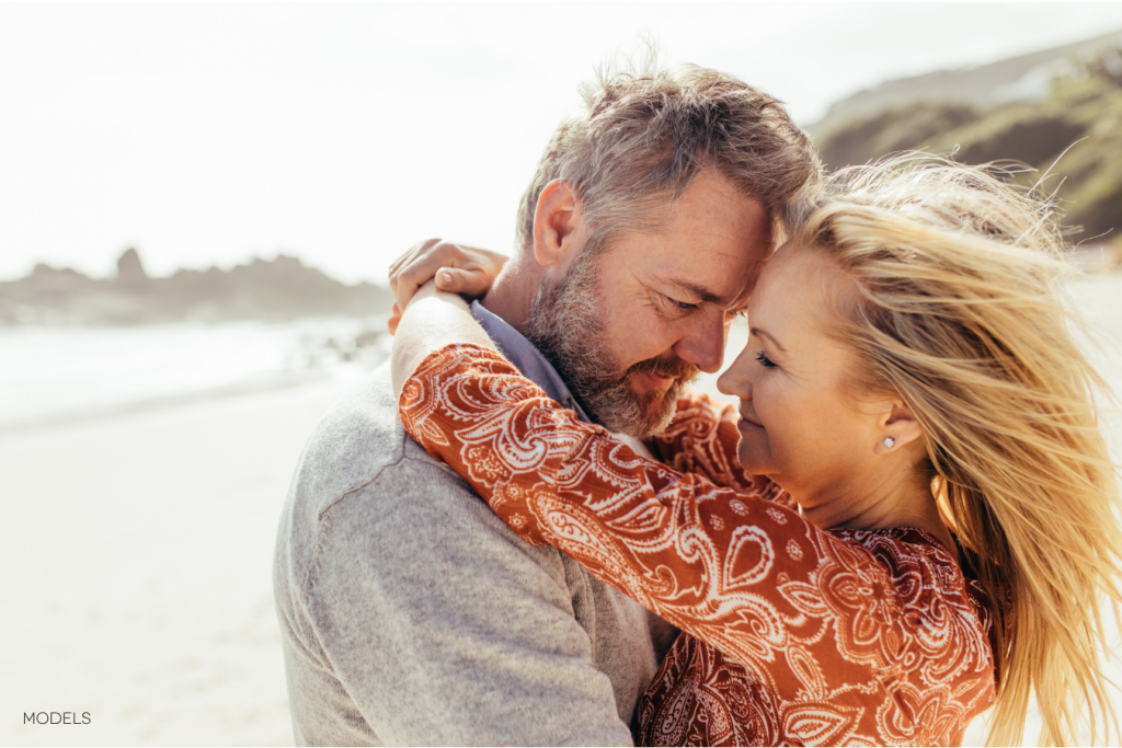 Happy middle aged couple, foreheads touching, arms around each other outside on a beach